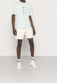 Miętowo zielony, oversize'owy t-shirt z nadrukiem z przodu, zestawiony z beżowymi szortami. Białe sneakersy i paskowane skarpetki dopełniają strój.