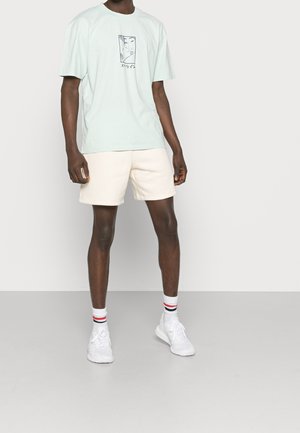 Miętowo zielony, oversize'owy t-shirt z nadrukiem z przodu, zestawiony z beżowymi szortami. Białe sneakersy i paskowane skarpetki dopełniają strój.