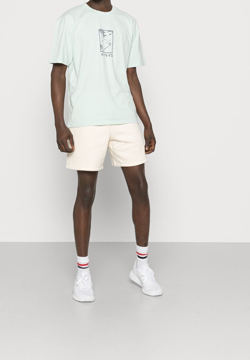 Miętowo zielony, oversize'owy t-shirt z nadrukiem z przodu, zestawiony z beżowymi szortami. Białe sneakersy i paskowane skarpetki dopełniają strój.