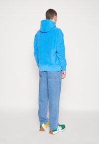 Felpa turchese con cappuccio e lacci, abbinata a jeans blu larghi e sneakers multicolori con dettagli verdi, gialli e bianchi.
