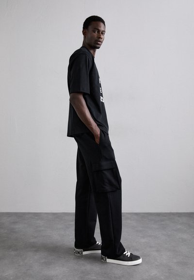 Tricou negru din bumbac cu imprimeu grafic, asociat cu pantaloni cargo negri cu buzunare laterale. Tenisi negri cu accente albe.