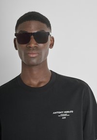Camiseta de algodón negra con cuello redondo, que presenta una impresión del logo blanco en el lado izquierdo del pecho. Modelo usando gafas de sol oscuras con montura angular.