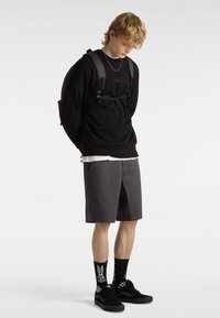Sudadera negra con el logo "vans.", pantalones cortos grises, zapatillas negras y calcetines con un diseño de conejo blanco; tirantes ajustables en la mochila.