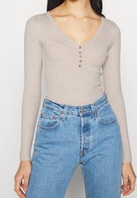 Nainen pukeutuneena beigeen ribbineulottuun, pitkähihainen V-kauluksiseen toppiin, jossa on viisi nappia, ja vyötäröä korostaviin vaaleansinisiin farkkuihin, seisoo valkoista taustaa vasten.