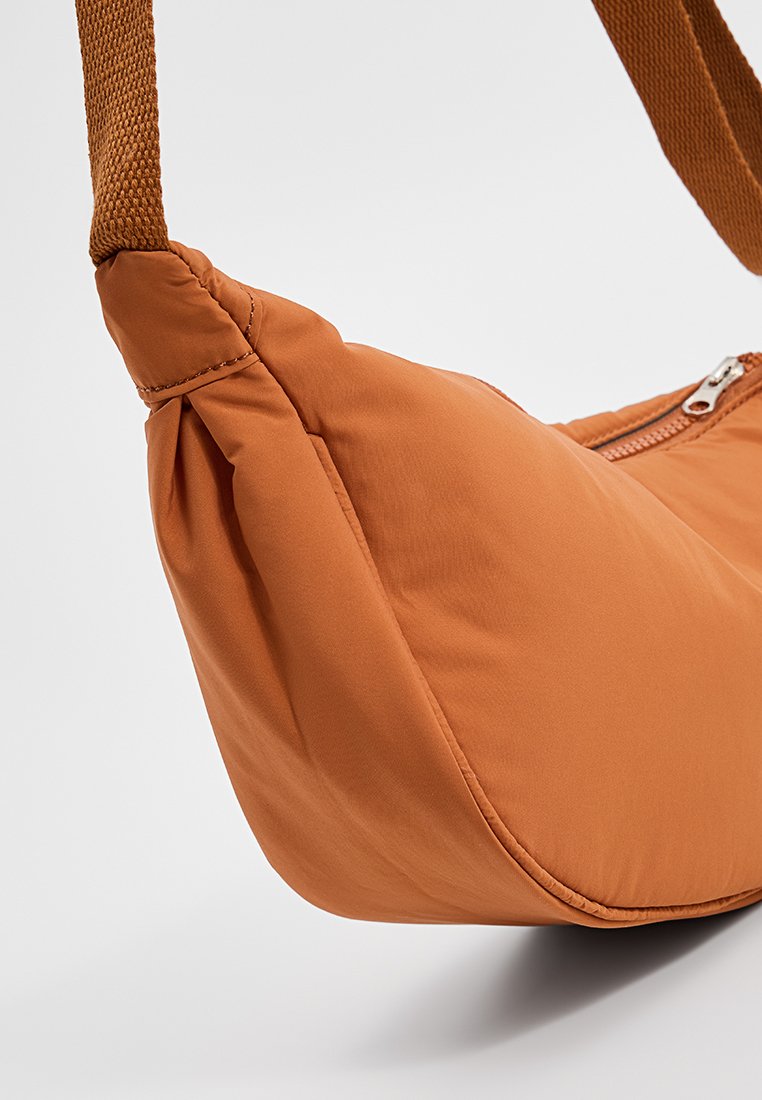 Sac rembourré orange à la texture douce, avec une base plate, une fermeture éclair et une sangle tissée. Le design est compact et arrondi.