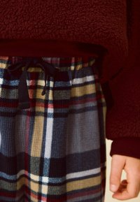 Burgunderfärgad fleece-tröja med en texturerad yta ovanför blå och rutiga loungebyxor med röda, gula och gröna rutmönster. Svart dragsko.