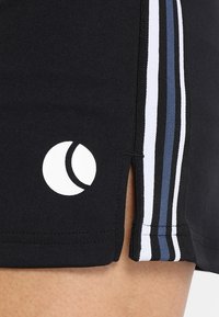Pantaloni sportivi neri con un logo circolare bianco, caratterizzati da fessure laterali e tre strisce verticali bianche e blu sui lati.