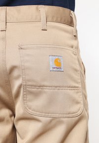 Beige werkbroek gemaakt van duurzame katoenen stof, met een achterzak en een vierkante logolabel in geel en grijs.