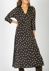 Kobieta w czarnej sukience midi w białe kropki, z rękawami o długości 3/4 i dekoltem w szpic, pozująca z jedną ręką na biodrze.