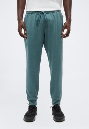 Mężczyzna ubrany w turkusowe spodnie dresowe z troczkiem i czarne trampki, stojący na jednolitym jasnym tle.