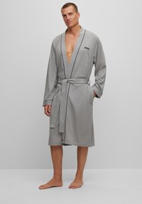 Accappatoio in cotone grigio con rifiniture nere, design avvolgente, maniche lunghe, cintura da annodare e tasche anteriori. Il modello è in piedi a piedi nudi su uno sfondo semplice.