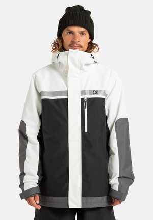 Giacca impermeabile bianca e nera con accenti grigi, tasche anteriori con zip, colletto alto e polsini regolabili. Presenta una striscia riflettente.