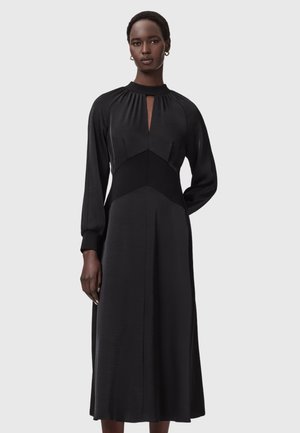 Kobieta stoi, ubrana w długą czarną sukienkę z długimi rękawami, wysokim kołnierzem i wycięciem z przodu w kształcie klucza, na tle jednolitego szarego tła.