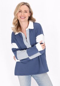 Polo a maniche lunghe blu navy con dettagli bianchi e strisce, dotato di colletto con bottoni e vestibilità rilassata, abbinato a jeans di denim chiaro.