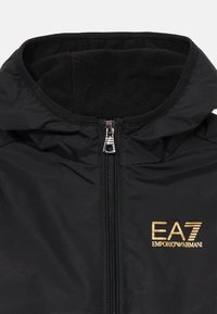 Svart hettejakke med fleece-fôret innerfor. Har hette, gull EA7-logo og glatt ytre materiale. Reflekterende glidelåshardware inkludert.