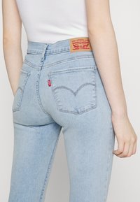 Vaqueros de tiro alto en azul claro, hechos de denim, con un parche de marca de cuero marrón y costuras curvas distintivas en los bolsillos traseros.