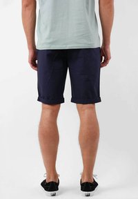 Shorts bleu marine avec revers, fabriqués en tissu léger. Détails de couture visibles et poches plates à l'avant. Chaussures noires avec semelles blanches.