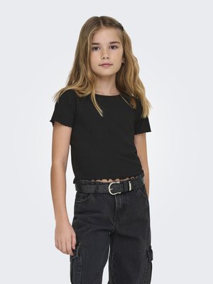 Fiatal lány hosszú szőke hajjal, fekete rövid ujjú felsőt és fekete cargo nadrágot fekete övvel viselve, semleges háttér előtt áll.