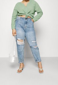 Top lungo verde a maniche lunghe con nodo in vita, jeans azzurri chiari a vita alta strappati, borsa bianca e sandali con tacco verde menta.