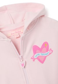 Sudadera de cremallera en rosa claro con un bordado en forma de corazón y una cinta que dice "Terapia del Amor" y un tirador de cremallera en forma de corazón.
