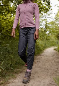 Fleecová bunda světle růžové barvy na zip, doplněná tmavě šedými kalhotami a pevnými hnědými turistickými botami, na zelené lesní stezce.