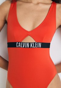 Červené jednodílné plavky s hlubokým výstřihem ve tvaru V a výřezem. Obsahuje černý pás s nápisem "CALVIN KLEIN" vytištěným bílou barvou. Hladká textura.