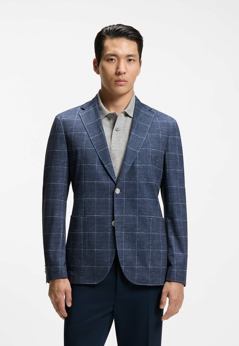 Homme portant un blazer à carreaux bleu windowpane sur un polo gris clair, debout, les mains détendues le long du corps.