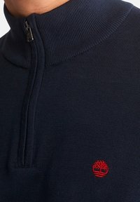 Suéter azul marino con cremallera y cuello alto, textura de punto y un pequeño logotipo de árbol rojo en el lado izquierdo del pecho. Detalle de cremallera en el cuello.