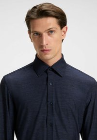 Camicia a maniche lunghe blu scuro con tessuto texturizzato, colletto ampio e chiusura con bottoni. Presenta bottoni blu lungo la pattina.