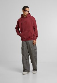 Haut à capuche rayé bordeaux avec une poche kangourou, associé à un pantalon cargo gris comportant plusieurs poches. Modèle portant des lunettes de soleil noires.