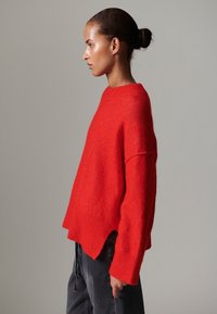 Röd oversize tröja i mjukt material med ribbad halsringning och fåll. Designen har nedsänkta axlar och en avslappnad passform.