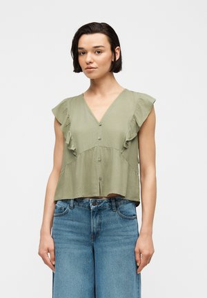 Kobieta ubrana w bezrękawnikową, jasnozieloną bluzkę z falbankami i guzikami, w połączeniu z wysokimi, niebieskimi dżinsami na jednolitym białym tle.