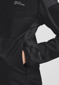 Giacca nera in tessuto morbido con tasca con zip, polsini elasticizzati e logo "Texadri" sulla manica. Progettata per l'uso all'aperto.