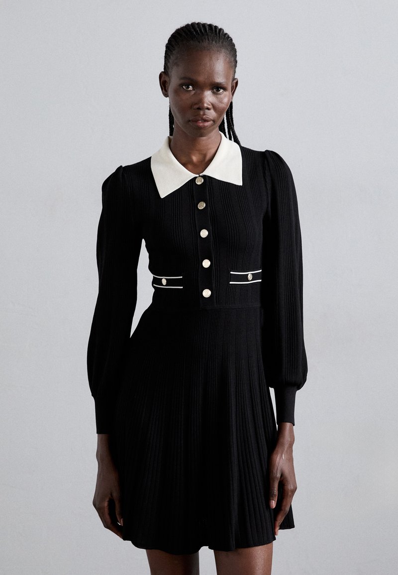 Vestido de punto negro acanalado con un cuello blanco, botones frontales y mangas abullonadas con acentos de piping blanco. Diseño de falda acampanada.