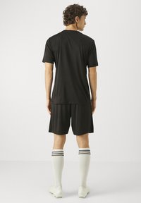 Férfi áll, fekete sportpólót és rövidnadrágot visel, fehér térdzoknit fekete csíkokkal, valamint fehér futballcipőt, hátulról látható egyszínű háttéren.