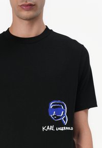Black cotton t-shirt featuring a blue and white graphic of a person with sunglasses and the text "KARL LAGERFELD" beneath.
