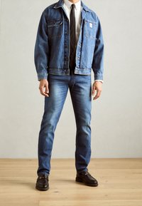 Giacca di jeans blu chiaro con due tasche frontali, chiusura a bottoni, abbinata a jeans blu scuro e mocassini neri su un pavimento in legno.