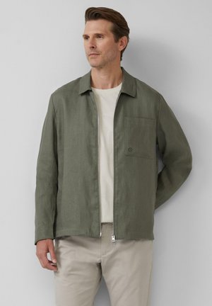 Groene, lichte jas met een kraag en een ritssluiting aan de voorkant, met een borstzak aan de linkerkant en een gladde textuur. Draagt over een beige shirt.