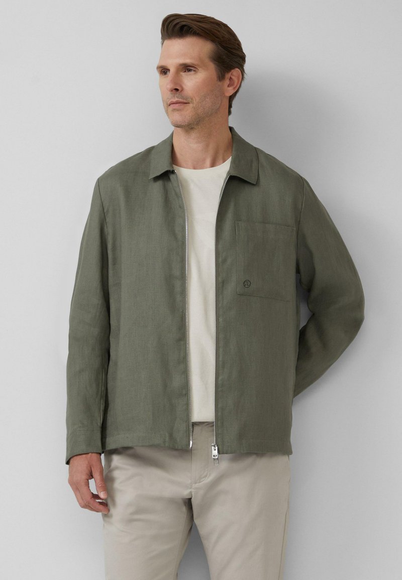 Groene, lichte jas met een kraag en een ritssluiting aan de voorkant, met een borstzak aan de linkerkant en een gladde textuur. Draagt over een beige shirt.