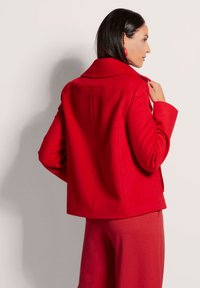 Rote Wollmischjacke mit Kragen, lockerer Passform und glatter Textur, von hinten vor einem neutralen Hintergrund gezeigt.