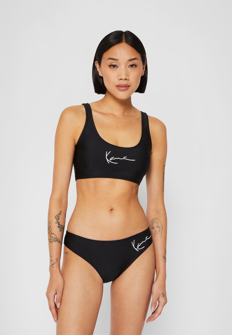 Set di bikini nero con top dallo stile sportivo, spalline larghe e scollatura a barca, abbinato a slip a vita bassa, entrambi con dettagli del logo bianco.