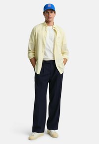 Geel gestreepte button-up shirt over een wit T-shirt, marineblauwe broek en blauwe pet. Witte sneakers met beige accenten. Casual pasvorm.