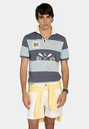 Młody mężczyzna w niebieskiej koszulce polo w paski, białych szortach i żółtym swetrze związanym wokół talii, stojący z neutralnym wyrazem twarzy.