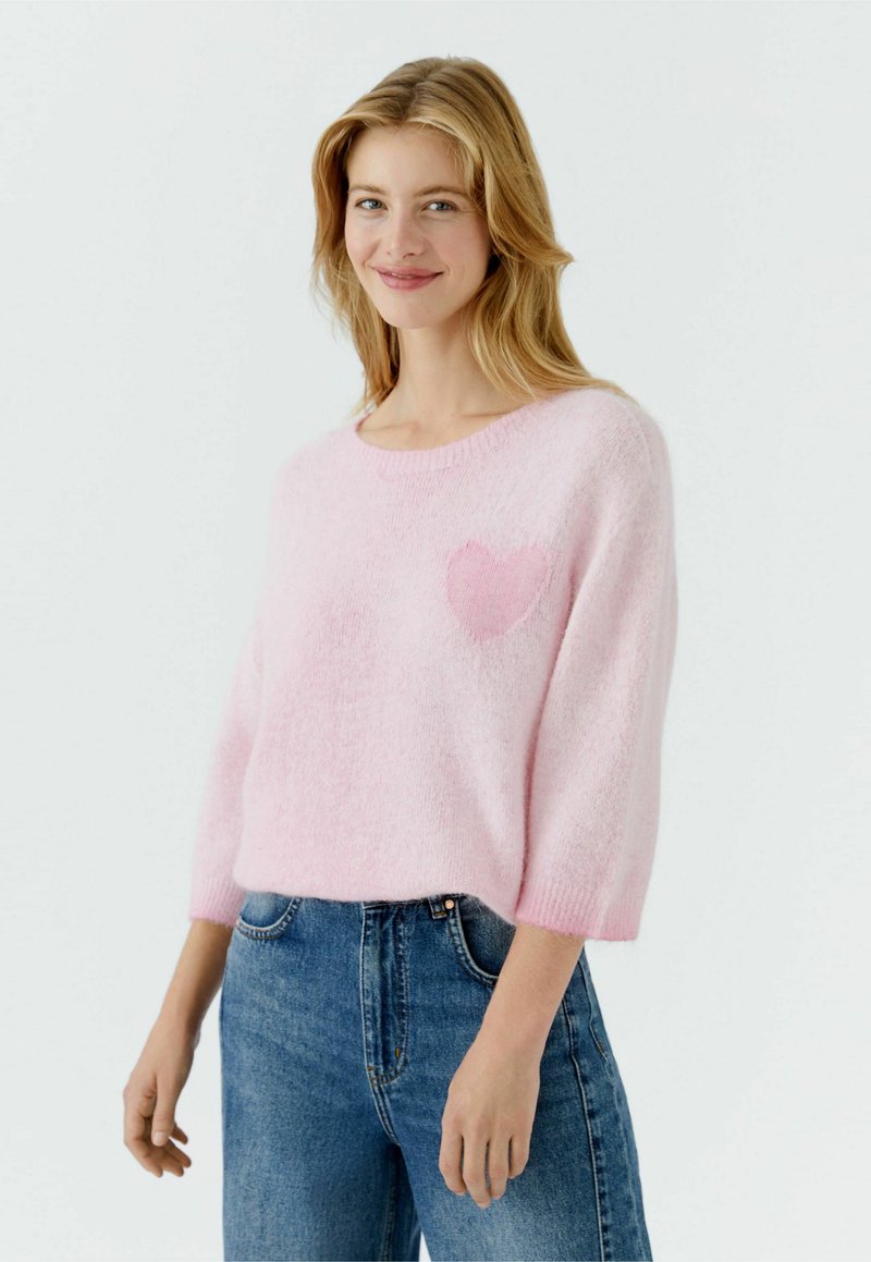 Jeune femme aux cheveux blonds portant un pull rose clair à motif de cœur et un jean bleu, souriant doucement devant un fond uni.