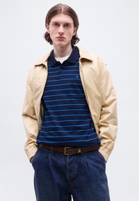 Νεαρός άνδρας φοράει μπεζ μπουφάν πάνω από μπλε-σκούρο και ανοιχτό μπλε ριγωτό πόλο πουκάμισο, σκούρα τζιν, στέκεται με τα χέρια στις τσέπες μπροστά σε απλό φόντο.