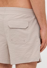 Shorts beige clair en tissu texturé, avec une poche arrière à rabat, des fentes sur les côtés et une taille élastique.