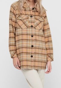 Veste-chemise à carreaux marron et beige fabriquée en tissu texturé, dotée de deux poches poitrine et de boutons noirs. Manches longues.