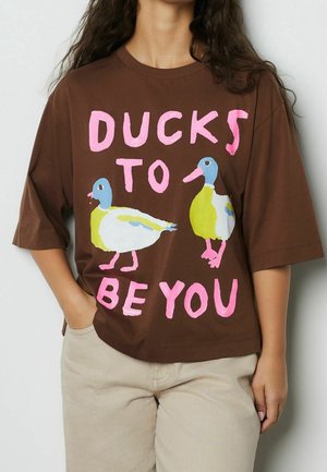 Chemise marron avec texte rose "Ducks to be you" et illustrations de deux canards colorés, portée par une personne avec une main dans la poche.