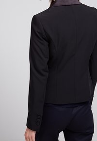 Svart skräddarsydd kavaj med satin-krage, långa ärmar och treknappsstängning vid ärmsluten. Den figursydda designen framhäver silhuetten.