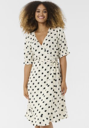 Cream wrap dress with black polka dots, short sleeves, and a tie waist. Features a v-neckline and a ruffled hem. Smooth fabric texture.